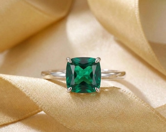 May include: A silver ring featuring a large, square-cut emerald gemstone. The vibrant green stone is set with four prongs, and the band is a polished silver. The ring is displayed on a gold-colored fabric, highlighting the gemstone's color.