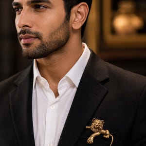 Peut inclure: Épinglette dorée en forme de lion sur une veste de costume noire. Le lion est détaillé avec une crinière et un corps texturés, et est positionné dans la poche de la veste. La personne porte une chemise blanche.