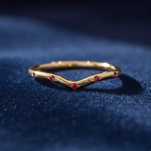 Op de afbeelding: Een goudkleurige ring met een V-vormig ontwerp, bezet met kleine, ronde, rode edelstenen. De ring ligt op een donkerblauwe ondergrond. De edelstenen zijn gelijkmatig verdeeld over de curve van de ring. De band van de ring is glad en gepolijst.