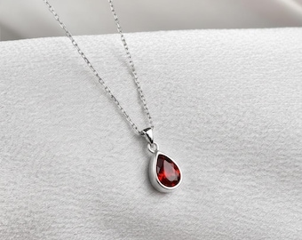 Può includere: Una collana d'argento con un pendente a forma di goccia. Il pendente presenta una gemma rossa vibrante, incastonata in una lunetta d'argento. La delicata catena pende su uno sfondo bianco strutturato. Gioielli.