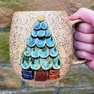 May include: A speckled beige ceramic mug featuring a raised, three-dimensional Christmas tree design. The tree is composed of overlapping blue-green glazed ceramic scales, topped with a yellow star, and flanked by two small gift boxes.