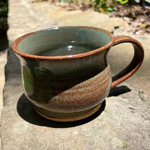 Könnte beinhalten: Eine grüne und braune Keramiktasse mit breitem Boden und abgerundetem Griff. Die Tasse ist mit einer hellbraunen Flüssigkeit gefüllt.