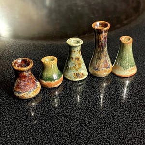 May include: Five miniature ceramic vases arranged in a row. Each vase features a unique glaze finish with varying shades of brown, green, and tan. The vases have a flared neck and a rounded base, displaying a glossy, mottled texture throughout.