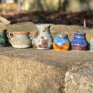 May include: A collection of six miniature ceramic vessels in various shapes and glazes. The set includes a green pot, a two-handled vessel, a speckled vase, and three smaller pots featuring blue, brown, and yellow tones with textured finishes.