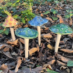 Könnte beinhalten: Vier Keramik-Pilzfiguren mit verschiedenen farbigen Glasuren. Die Pilze sind auf einem Bett aus braunen Blättern angeordnet. Die Pilze sind alle unterschiedlich groß und geformt.