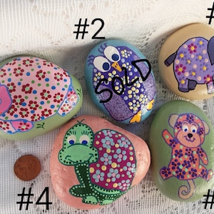 May include: Hand-painted decorative rocks featuring animal designs. The collection includes a hippo, owl, elephant, turtle, and monkey, each adorned with colorful floral patterns. The owl rock is marked as "SOLD". A penny is included for size reference.