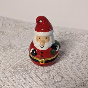 May include: A ceramic Santa Claus ornament with a red hat, white beard, and a black belt. He is holding a green bag in his right hand.