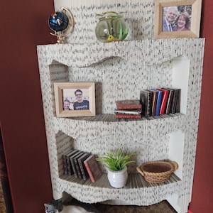 Peut inclure: Une sculpture de bibliothèque unique faite de pages de livres pliées, avec des objets décoratifs miniatures. Comprend un globe, des photos encadrées, de petits livres, une plante, un panier et une figurine de chat. Les étagères sont blanches avec du texte noir, sur fond rouge.