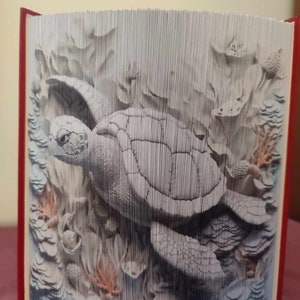 May include: A white papercraft book sculpture of a sea turtle swimming through a coral reef. The sculpture is made of folded paper and has a 3D effect.