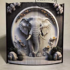 Peut inclure: Une sculpture d'éléphant blanc réalisée à partir de pages de livres pliées, sur fond noir. L'éléphant est entouré de fleurs et de feuilles, et la sculpture est encadrée par une bordure circulaire. Le texte "Book Art By Davida" est visible en haut de l'image.