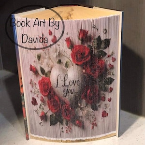 May include: A book art sculpture featuring a heart with the words "I Love You" surrounded by red roses and small hearts. The book is folded into a three-dimensional shape and is titled "Book Art By Davida".