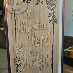 Pode incluir: Uma peça de arte de livro artesanal com o texto "The joy of the Lord is my strength" cortado nas páginas. A arte do livro apresenta desenhos florais e de folhas nos cantos e o texto "Book Art By Davida".