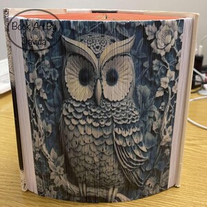 May include: A book art sculpture of an owl with blue and white feathers. The owl is perched on a branch with leaves and flowers. The book is folded into a three-dimensional shape. The text "Book Art By Davida" is visible on the front cover.