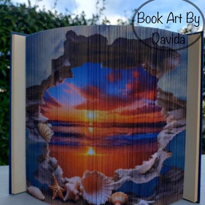 May include: A book art sculpture of a sunset over the ocean with seashells in the foreground. The book is folded to create a 3D effect. The text "Book Art By Davida" is visible in the upper right corner of the image.