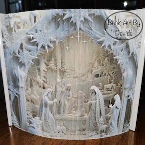 Può includere: Una scultura di arte di libro di carta bianca che rappresenta la scena della natività. La scultura è piegata in forma 3D e presenta una stella sopra la mangiatoia. Il testo "Book Art By Davida" è stampato sul lato destro della scultura.