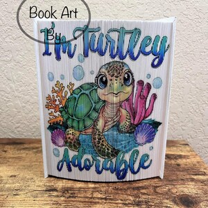 May include: A folded book art sculpture of a turtle with the text "I'm Turtley Adorable" on the front. The turtle is green with brown spots and has blue eyes. The background is blue and white with coral and seashells.