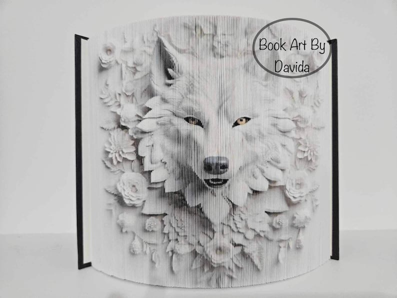 May include: A white wolf's face is folded out of the pages of a book, surrounded by white flowers. The book is closed and standing upright. The text "Book Art By Davida" is printed on the spine of the book.