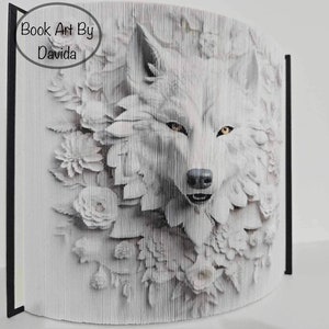 May include: A white wolf's face is created using folded pages of a book. The wolf's face is surrounded by white flowers. The book is titled "Book Art By Davida".