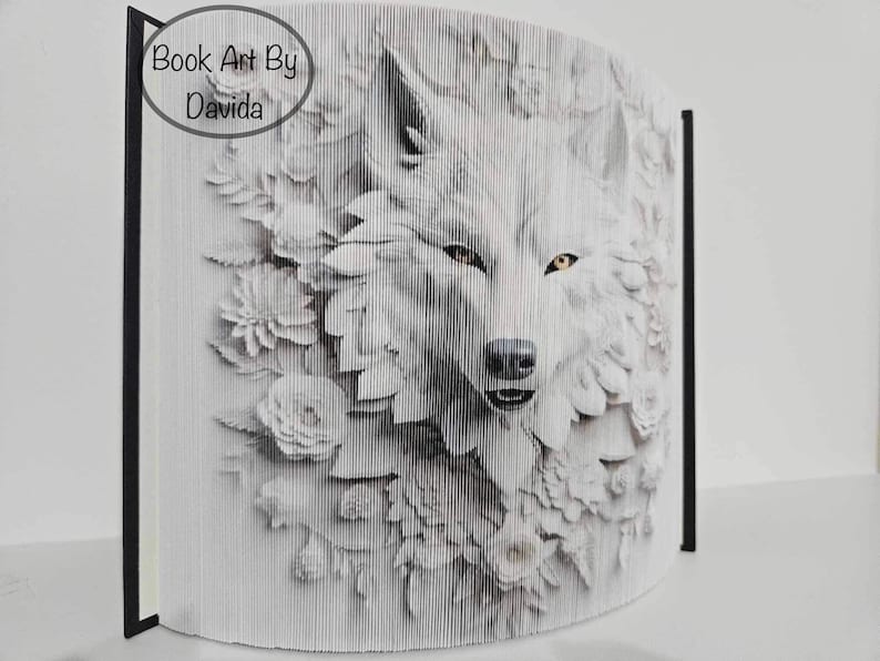 May include: A white wolf's face is created using folded pages of a book. The wolf's face is surrounded by white flowers. The book is titled "Book Art By Davida".