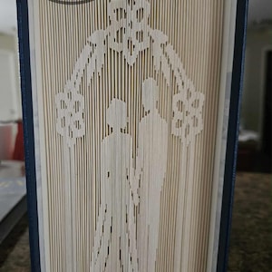 Puede incluir: Una obra de arte en libro con un diseño tallado de una pareja bajo una estructura ornamentada. Las páginas son beige, con una encuadernación azul oscuro. El texto "Book Art By Davida" está en un marco circular en la parte superior.
