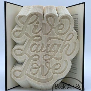 以下が含まれることがあります： ページに「Live Laugh Love」の文字が彫られた本の彫刻。 本は開かれており、筆記体で彫られたテキストが明らかになっています。 本のページはベージュで、表紙は黒です。