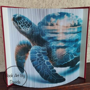 May include: A blue and green sea turtle book art sculpture with a sunset and ocean scene in the background. The book art is titled "Book Art By Davida".