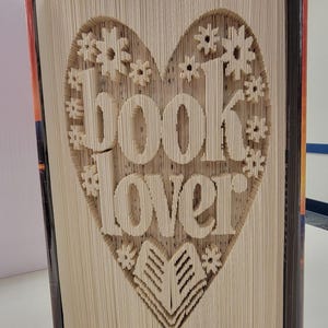 Puede incluir: Una escultura de libro plegado con el texto book lover tallado en las páginas. Las hojas están dobladas de forma intrincada para formar una gran figura de corazón que rodea las palabras, con pequeños detalles de estrellas y un libro abierto.