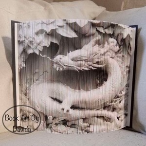 May include: A white paper dragon sculpture made from a book. The dragon is curled up in a defensive position with its head raised and its wings spread out. The sculpture is made from a book that has been folded and cut to create the dragon's shape. The book is open and the pages are visible. The sculpture is on a white surface. "Book Art By Davida" is written on the bottom left corner of the sculpture.