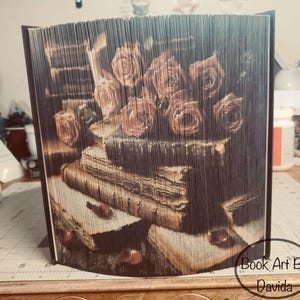 Può includere: Una scultura di arte libraria con una pila di vecchi libri sormontata da un mazzo di rose. La scultura è realizzata con pagine di un libro che sono state piegate e sovrapposte per creare un effetto tridimensionale. La scultura è firmata "Book Art By Davida".