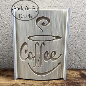 Op de afbeelding: Een uitgesneden boekkunstwerk met het woord "Coffee" en een koffiekopje-ontwerp. De pagina's van het boek zijn uitgesneden om de afbeelding te creëren. De tekst "Book Art By Davida" staat in een cirkel linksboven. Het boek staat op een houten oppervlak.