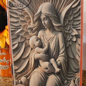 May include: A book art sculpture of an angel with wings holding a baby. The sculpture is made of folded pages of a book and is painted in shades of brown and white.