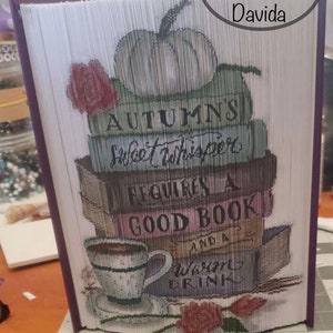 Può includere: Un'opera d'arte di libro con un tema autunnale. Il libro è piegato per creare un'immagine 3D di una pila di libri con una zucca, una tazza di caffè e una rosa. Il testo sui libri dice "Autumn's Sweet Whisper Requires a Good Book and a Warm Drink".
