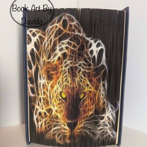 May include: A book art sculpture of a leopard with glowing yellow eyes. The sculpture is made of folded pages of a book and is titled "Book Art By Davida".