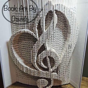 May include: A book art sculpture of a treble clef made from the pages of a book. The sculpture is standing upright on a wooden surface. The book is open and the pages are folded to create the shape of the treble clef. The title "Book Art By Davida" is visible on the front cover of the book.