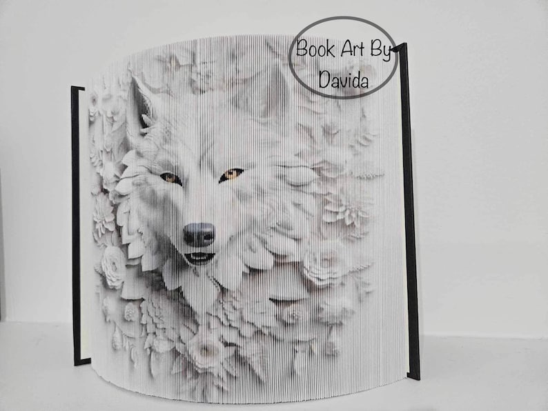 May include: A white wolf's face is partially obscured by white paper flowers. The wolf's eyes are yellow. The image is a book art piece by Davida.