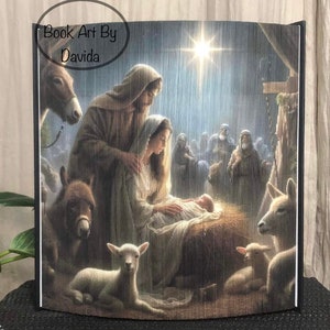 May include: A folded book art piece depicting the Nativity scene. The image shows Mary, Joseph, and baby Jesus in a manger, surrounded by animals and onlookers. The text "Book Art By Davida" is visible.