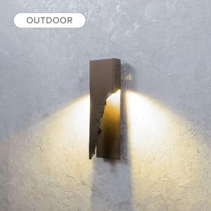 May include: A rectangular dark bronze outdoor wall sconce with a jagged, textured vertical cutout edge. The fixture emits a warm, bright light from behind the metal panel, casting a wide, fan-shaped glow onto the surrounding wall surface.