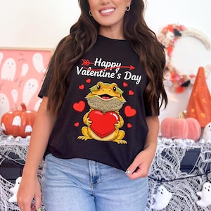Peut inclure: T-shirt noir sur le thème de la Saint-Valentin. Le t-shirt présente un lézard de dessin animé tenant un cœur rouge, entouré de cœurs rouges, et le texte "Happy Valentine's Day". Le t-shirt est porté par une personne.
