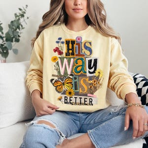 May include: A light yellow sweatshirt with the text "His Way Is Better" in a collage style. The design includes a butterfly, flowers, and various fonts. The sweatshirt is long-sleeved and worn with blue jeans.
