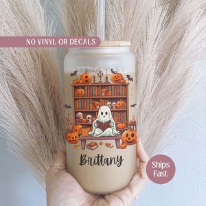 May include: A frosted glass tumbler with a Halloween-themed design featuring a ghost reading a book in front of a bookshelf with pumpkins and bats. The tumbler has the name "Brittany" printed on the bottom.