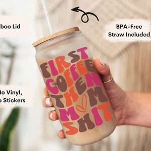 Op de afbeelding: Een herbruikbare glazen koffiebeker met een bamboe deksel en een rietje. De beker heeft een roze, bruine en oranje afbeelding met de tekst "First Coffee Then Mom Shit".