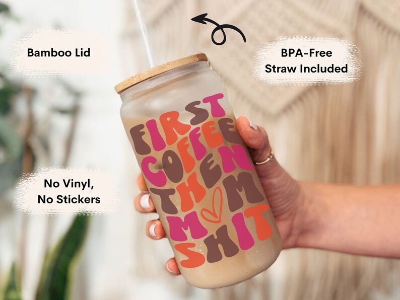 Op de afbeelding: Een herbruikbare glazen koffiebeker met een bamboe deksel en een rietje. De beker heeft een roze, bruine en oranje afbeelding met de tekst "First Coffee Then Mom Shit".