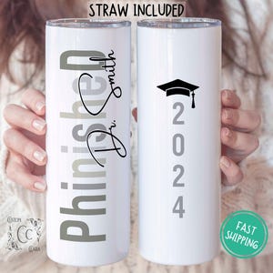 May include: Two white stainless steel tumblers with a graduation theme. One tumbler has the text "Finished" with the name "Tri Smith" in cursive. The other tumbler has a graduation cap and the year "2024" in gray.