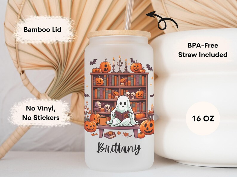 May include: A white 16 ounce reusable tumbler with a bamboo lid and a straw. The tumbler features a ghost reading a book in front of a bookshelf with pumpkins and candles. The text "Brittany" is printed on the bottom of the tumbler.