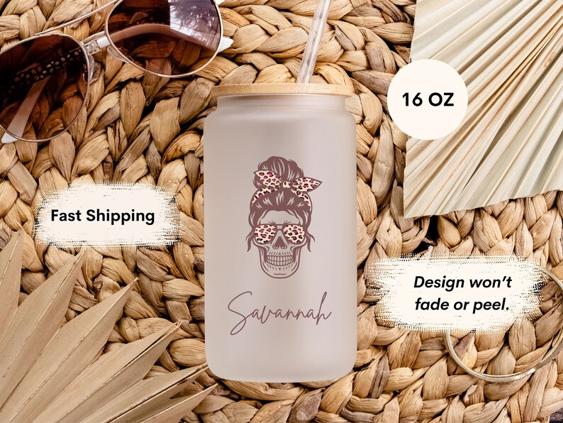 Op de afbeelding: Een 473 ml frosted glazen tumbler met een bamboe deksel en een rietje. De tumbler heeft een ontwerp van een schedel met een knot, die een zonnebril met luipaardprint en een hoofdband draagt. De naam "Savannah" staat onder de schedel. Het ontwerp is op de tumbler gedrukt en zal niet vervagen of afbladderen.