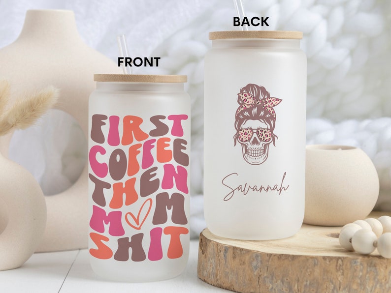 Op de afbeelding: Een witte, gematteerde glazen tumbler met een bamboe deksel en rietje. De voorkant van de tumbler heeft de tekst "First Coffee Then Mom Shit" in roze, oranje en bruin. De achterkant van de tumbler heeft een roze schedel met een luipaardprint bandana en zonnebril. De tekst "Savannah" staat onder de schedel.
