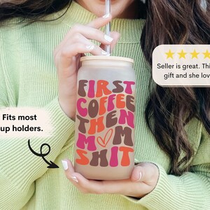 Op de afbeelding: Een frosted glazen beker met een roze, bruine en oranje afbeelding met de tekst "First Coffee Then Mom Shit". De beker heeft een deksel en een rietje. Het past in de meeste bekerhouders.