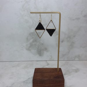 Pode incluir: Um par de brincos de contas pretas com um design geométrico, exibidos em um suporte de metal dourado com uma base de madeira. Os brincos têm formato de diamante.