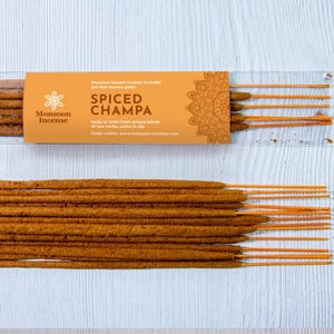 Pode incluir: Duas embalagens de varetas de incenso. Uma embalagem está num recipiente de plástico transparente, e a outra é uma pilha de varetas de incenso. A embalagem é laranja com as palavras "Spiced Champa" e "Monsoon Incense".