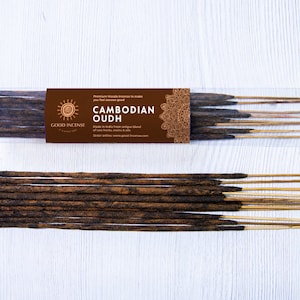 May include: Two packages of Cambodian Oudh incense sticks. The incense sticks are dark brown and arranged in bundles. The packaging includes a brown label with the words "GOOD INCENSE" and "CAMBODIAN OUDH". The background is a white wooden surface.